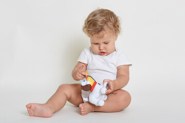 Charming beautiful baby boy playing with plastic dog, concentrated male kid holding new toy in hand and study it attentively, toddler wearing white bodysuit sitting on floor.