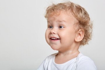 Little baby boy with curly blond hair laughing while looking attentively away wile posing isolated over white background, excited male child sees something interesting, copy space for promo.