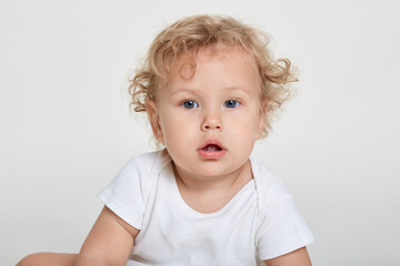 Curious baby looking directly at camera, showing his two teeth, wearing t shirt, posing isolated over white background, looks with interest at world around.