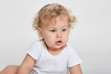 Naklejka premium Little cute child sitting with excited facial expression against white, kid wearing bodysuit, infant looking away with interest, blond baby boy with blond curly hair.