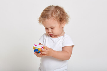 Blond toddlers playing with color car isolated on white background, looking concentrated at toy in his hands, wearing t shirt, male child with curly hair.