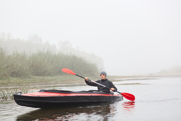Fototapeta premium Sportsman in kayak, padding in foggy river, spending his free time outdoors, doing water sport, posing with oar in hands, rowing boat, looks straight ahead.