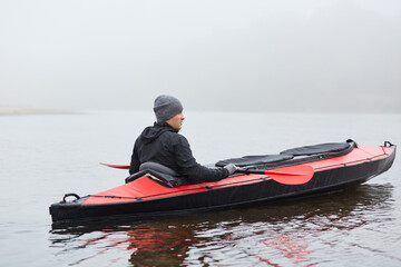 Back view of man paddling kayak on river or lake, looking at nature, wearing black jacket and gray cap, foggy day in open air, canoeing, water sport.