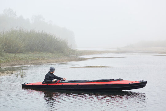 Side view of male kayaking on river, sportsman in black jacket looking at foggy water, holds oar in hands, foggy day, man in boat with bank of river and bushes on background.