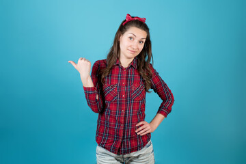 A country girl in a red plaid shirt smiles skeptically and points a finger to the side. Isolated on blue background.