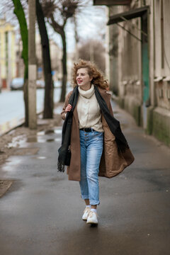 A beautiful young woman in a brown coat and curly hair runs down the street after the rain. Outside