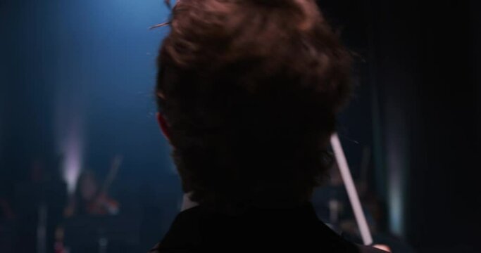 Cinematic close up shot of conductor directing symphony orchestra with performers playing violins, cello and trumpet on classic theatre with curtain stage during music concert with dramatic lights.
