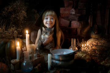 Cute little witch with a cat brews a potion over a fire of herbs and mushrooms. Beautiful girl in witch costume with Halloween decoration