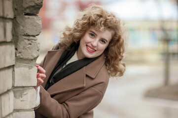 A beautiful laughing young woman with a smile peeks out from behind the wall. Outside.