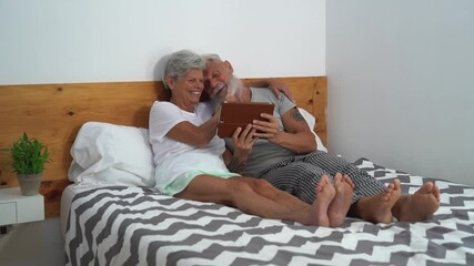 Happy senior couple using digital tablet in bed - Mature people having funny bed time together - Elderly love relationship lifestyle and technology concept  - Powered by Adobe