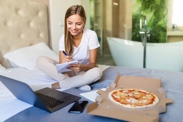 Young woman working on a bed in bedroom indoors at home while eating pizza and using laptop