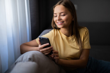 Happy woman using mobile phone applications, shopping, playing online games, chatting with friends, watching funny video sitting on comfortable coach in living room,