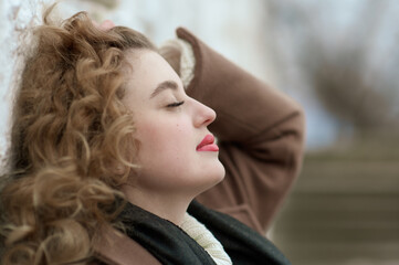 Naklejka premium Portrait of a beautiful young woman with closed eyes in a brown coat and a black scarf near a white stone wall. Outside. Close-up