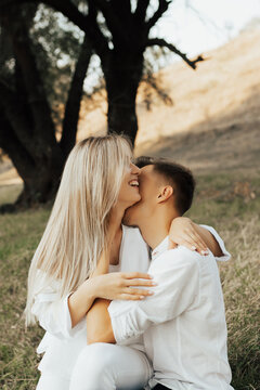 Charming caucasian couple in white clothes embracing, while woman smiling and man kissing her soft neck in the park.