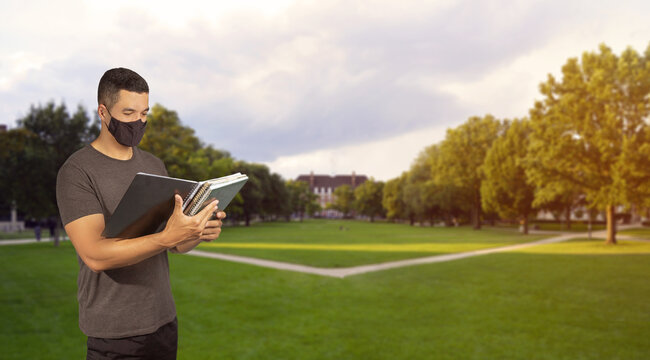 A Studant Man With Covid 19 Coronavirus Mask Holding A Copybook. In The Park