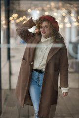 A beautiful girl in a red hat and coat stands behind a glass wall. Golden bokeh in the background