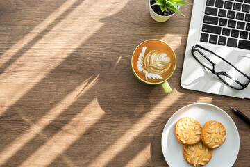 Wood office desk table with dish of cookies, cup of latte coffee and supplies. Top view with copy...