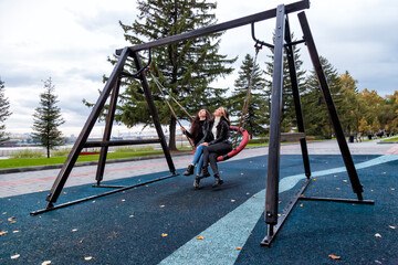 Two beautiful young women friends with long hair are sitting together on a round wicker swing in the park at the playground raising their heads up smiling in an autumn day.