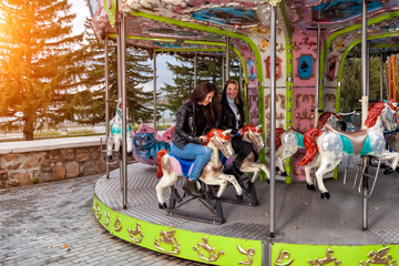 Two young women friends with long hair in jackets and jeans ride an amusement ride in the park on an autumn day on a round carousel riding horses and laugh. Retro style entertainment.