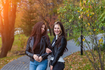 Two girlfriends, students laugh and stand on the path in jackets and jeans while walking in the park against the background of trees with yellow leaves in an autumn day.