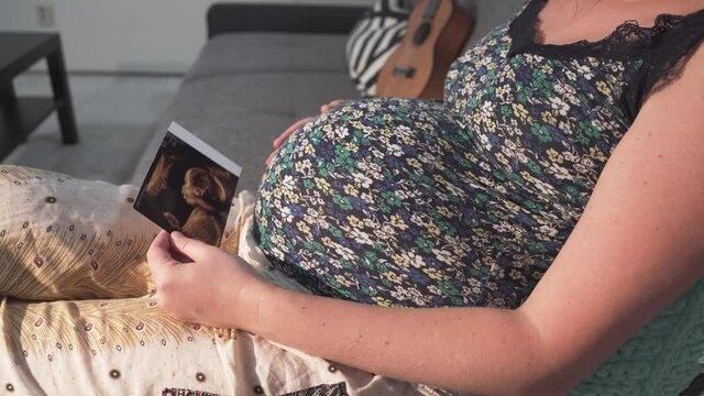 Pregnant girl is sitting on sofa, touching belly and watching ultrasound picture.