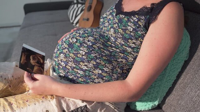 Pregnant girl is sitting on sofa, touching belly and watching ultrasound picture