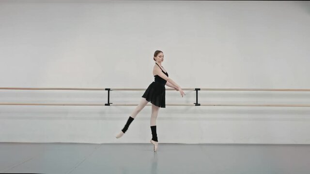 Ballerina in black dress and Pointe shoes is engaged near ballet machine. Female dancer performs exercises for rehearsing performances in dance studio. Slow motion.