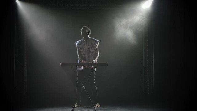Silhouette of afroamerican man playing piano synthesizer keyboard on stage in dark studio. A musician with a painted face in ethnic African style performs against the background of lights. Slow motion