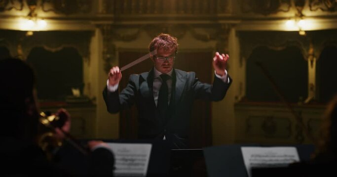 Cinematic close up shot of conductor directing symphony orchestra with performers playing violins, cello and trumpet on classic theatre with curtain stage during music concert with dramatic lights.