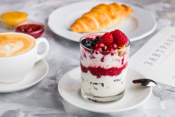 Breakfast - yogurt with berries (currants, raspberries, blueberries) and granola in a glass with a cup of coffee and a croissant
