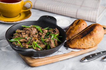 Fried meat in a frying pan on a wooden board on a light background with a cup of tea