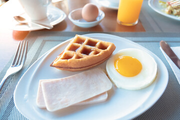 Lazy American breakfast in warm tone sunlight. Continental meal at hotel with fresh waffle, fried egg, ham, orange juice and cup of coffee, selective focus. Concept of travel bubble and rest a