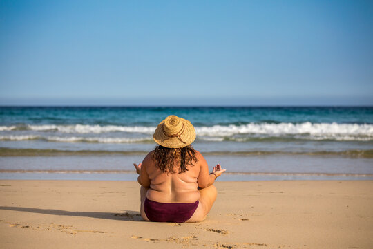 Mollige Frau mit Strohhut am Strand in Yoga-Pose