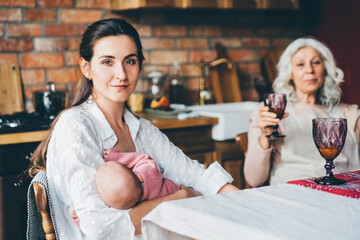 young woman breastfeeds baby and looking to the camera and sitting on wooden chair next to grey haired mother.