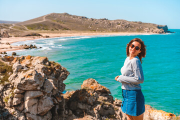 A happy woman with long hair sits on a rock with her back to the camera and admires the raging sea in the middle of the rocks