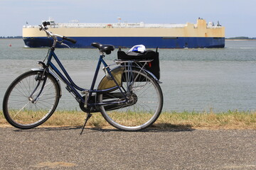 Fototapeta na wymiar a bicycle with a pannier with a towel stands at the seawall at the dutch coast in zeeland, and a roro cargo ship is passing by in summer