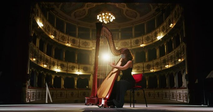 Cinematic shot of professional female harpist wearing protective medical mask is playing harp solo on a classic theater stage with dramatic lighting before start a music concert.