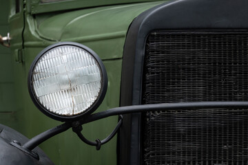 Fragment of the front of a green vintage truck. There is a headlamp with broken glass and part of a...