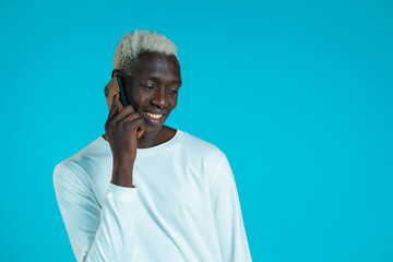 Young african american man speaks with smile on phone. Guy holding and using smart phone. Blue studio background.