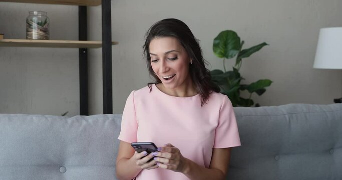 Relaxed young indian arab mixed race woman sitting on sofa, web surfing internet in smartphone application, feeling excited of getting shopping promo code in online store, winning prize in lottery.