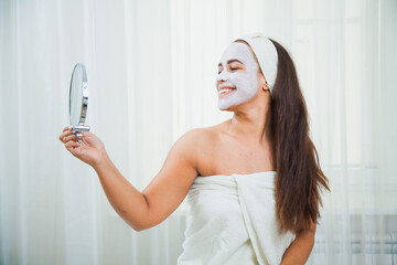 Happy woman in towel and facial mask look in the mirror. Home beauty treatment. Skincare and rejuvenation concept.