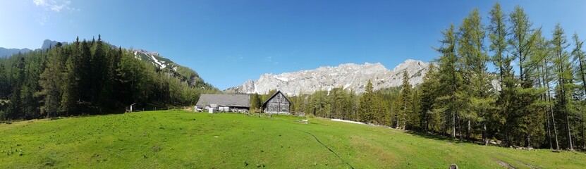 A panoramic view on a massive mountains in Hochturm region, Austrian Alps. There is a small cottage in the middle of lush green meadow and thick and dense forest in the back. Exploring the nature