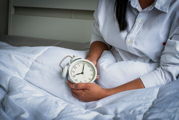 a young Asian woman sitting in bed holding the white alarm clock to set the alarm for the day...