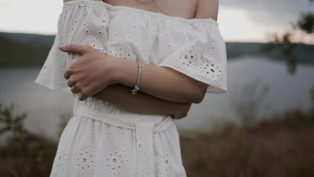 A girl with a silver bracelet in a white delicate dress with bare shoulders, arms crossed, stands against blurred dark sky, shore and river and freezes from cold wind that blows her clothes and hair
