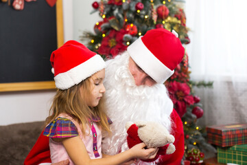 Little girl in Santa hat on Santa's lap near Christmas tree in Christmas decor. Shows a box with a gift, a soft toy, touches his beard and laughs. New year, Russian grandfather Frost