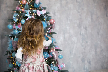 A little girl reaches for a Christmas tree toy with her hand. Christmas decor, waiting for a holiday and a miracle. New Year. White, pink and blue decor, Golden lights garlands