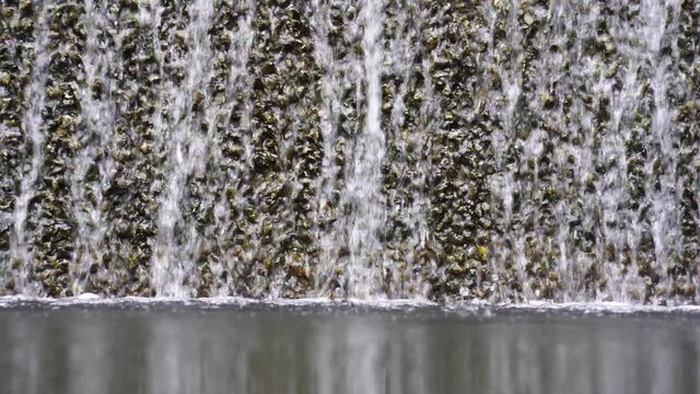Water falling pouring into a poolside from stone wall, close up. Background with running water. Waterfall fountain stri and spray drops stone surface.
