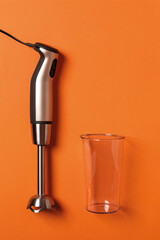 Immersion hand blender and empty cup on a carrot orange background. Modern electric kitchen appliances for making sauces, smoothies, puree. Copy space.