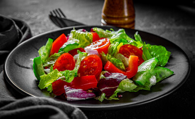 various fresh mix salad leaves with tomato, cucumber and red pepper on a black plate on Dark grey black slate background