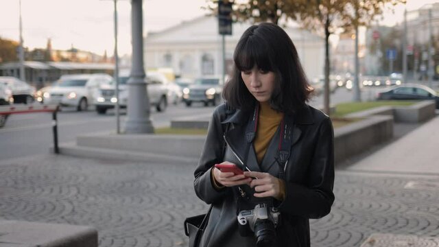 Young woman black hair with phone, walking on background of cars moving on the city road. Asian female with photo camera on a walk typing in phone, gimbal shot